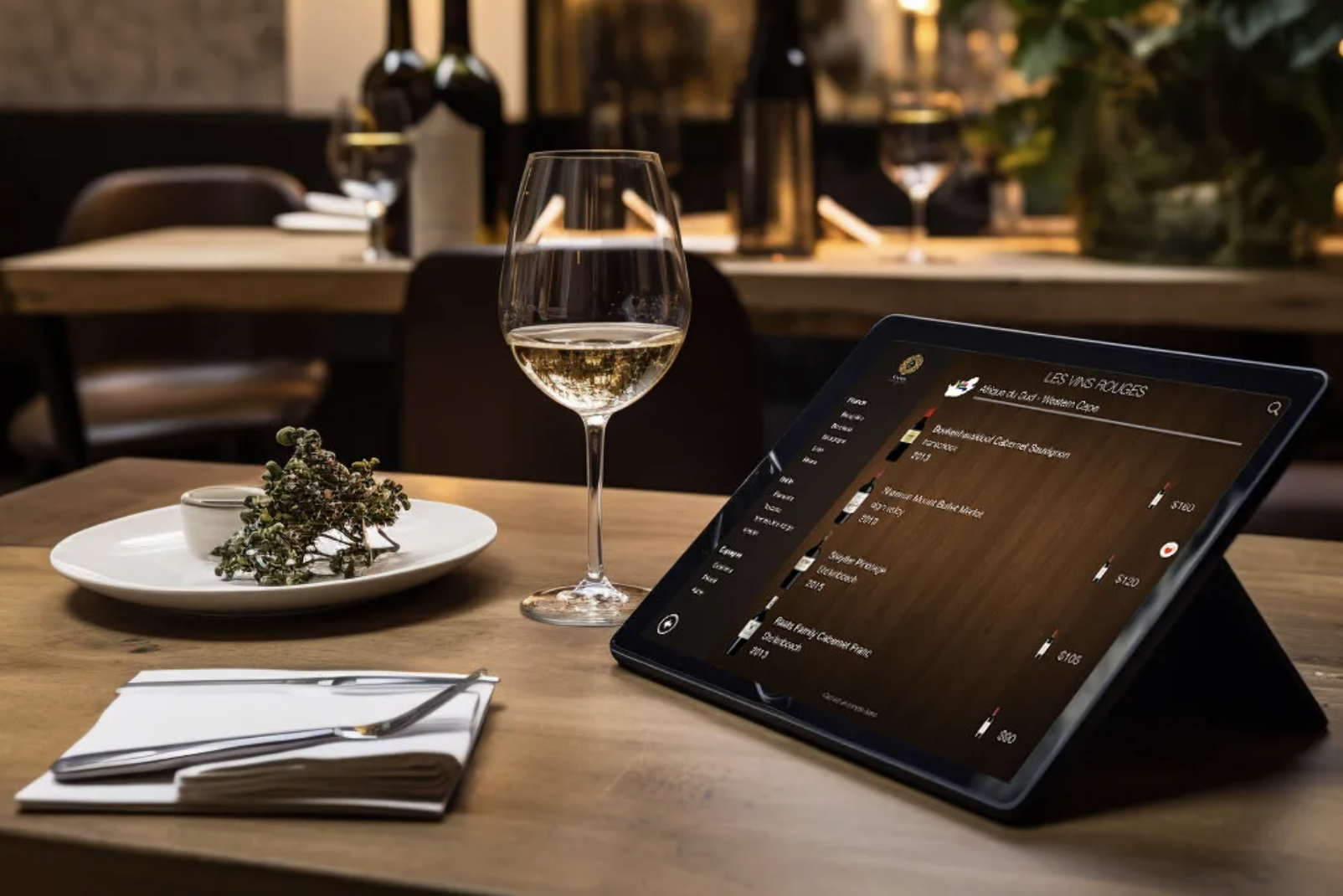 A digital wine menu tablet on a restaurant table next to a glass of white wine, showcasing modern wine technology